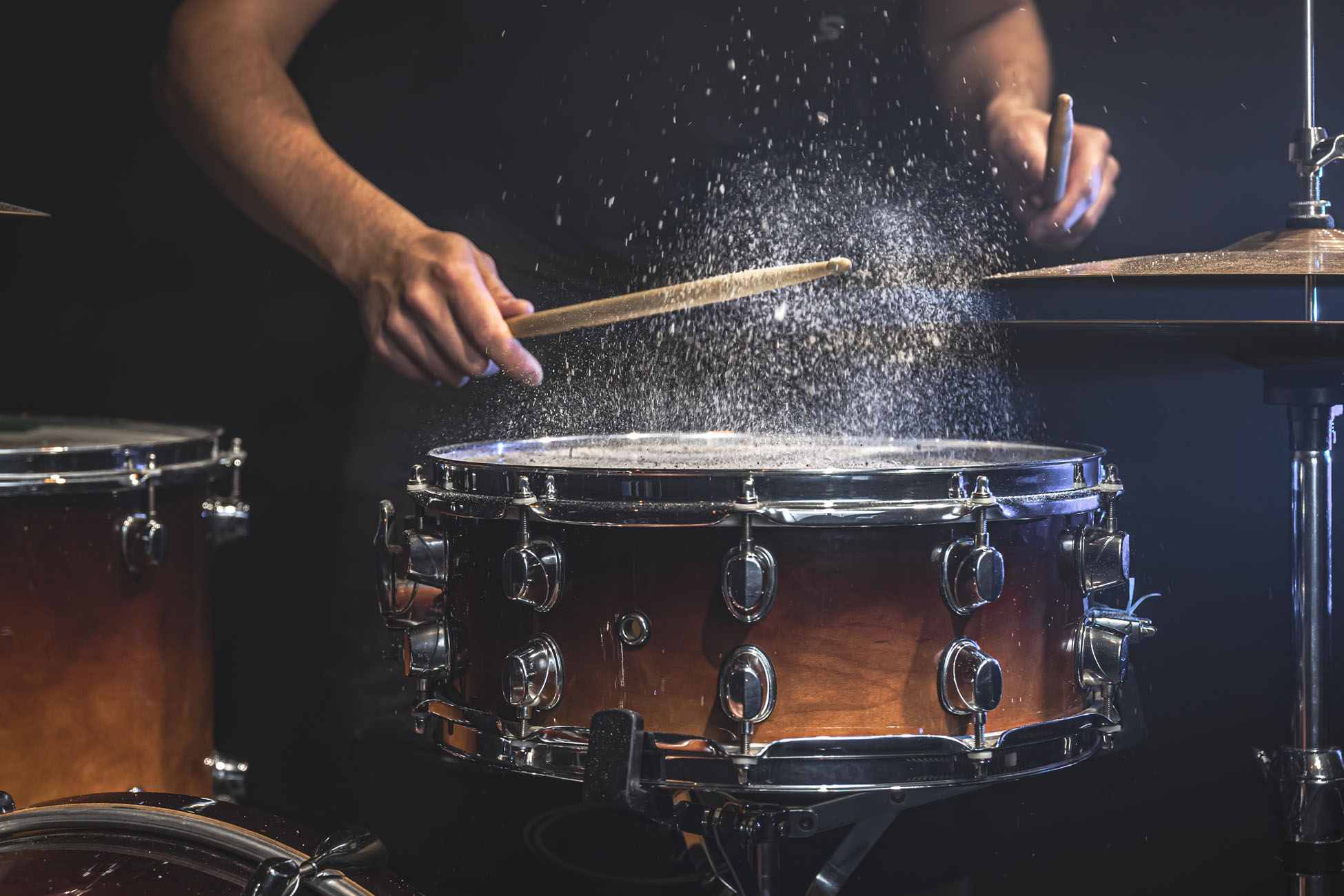 A male drummer plays snare drum with drumsticks in a dark room.